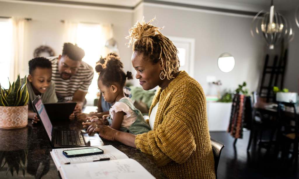 Photo of a woman sitting at a laptop smiling with her family in the background