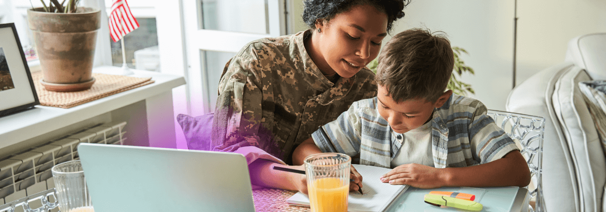 Mother and son sitting at a table drawing on a piece of paper with a laptop open in front of them. The mother is wearing a military uniform.