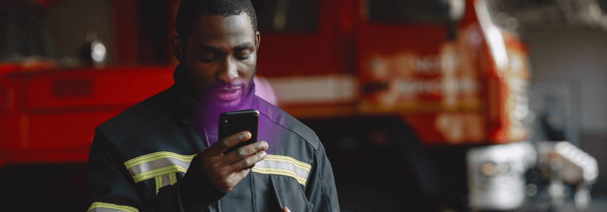 Young firefighter looking at a mobile phone with a purple glow coming from the screen. A firetruck is in the background.